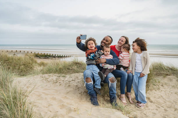 Vacationing British family posing for selfie at Camber Sands Full length front view of mid adult father photographing family aged 23 months to 6 years sitting on dune and smiling at smart phone with Rye Bay in background. sand dune photos stock pictures, royalty-free photos & images