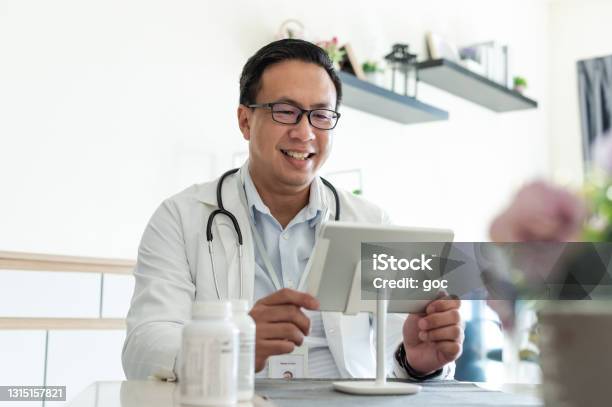 Asian Male Doctor Consulting And Consoling His Patient On Live Video Call At Hospital Clinic Stock Photo - Download Image Now