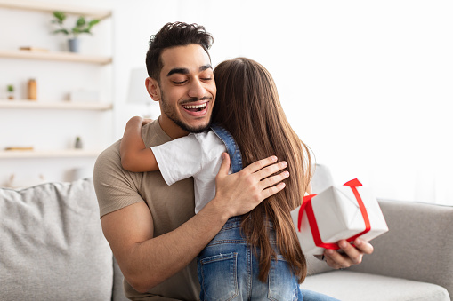 This Present Is For You. Portrait of loving girl with long hair hugging dad and greeting him with father's day or birthday, excited man holding wrapped gift box, sitting on couch at home