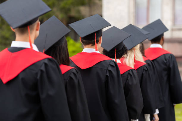 Back view of international group of students in graduation robes and caps walking by university campus, closeup. Unrecognizable multiracial students attending graduation ceremony