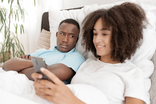 istock Young couple having problem. Jealous husband is watching his wife use mobile phone on bed at home 1315158314