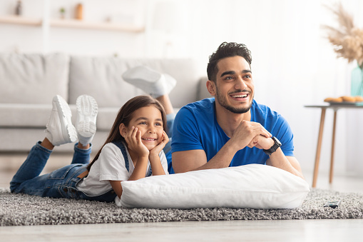 Portrait of happy young man and girl watching tv show or movie, lying on the floor carpet with pillows in living room at home, having fun. Excited father and daughter spending time together