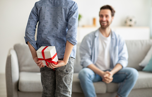 Boy greeting his young dad with Father's Day, hiding gift box behind his back, selective focus. Dad sitting on sofa and smiling to son. Focus on boy with present. Family holiday concept