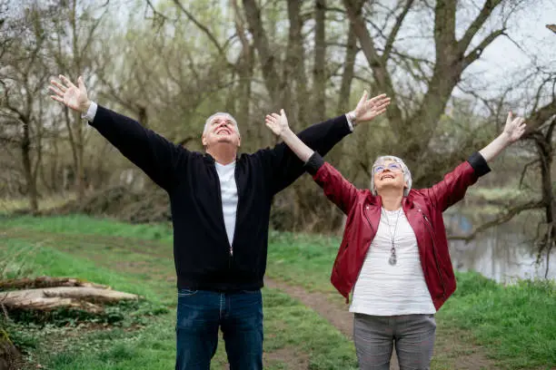 Elderly couple standing in nature spreading their arms wide open and looking up at the sky