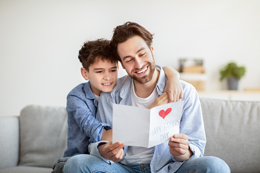 Father's day concept. Son congratulating daddy and giving him handmade postcard, dad reading card and smiling, sitting together on sofa. Family spending time on weekend at home