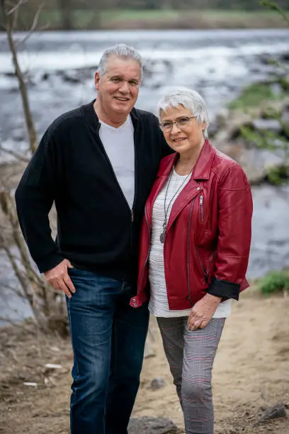 Elderly couple standing in front of a river, looking at the camera and smiling