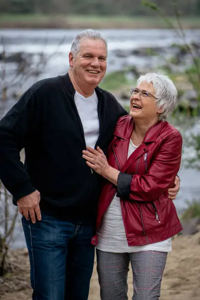 Elderly couple standing in front of a river, looking at the camera and smiling