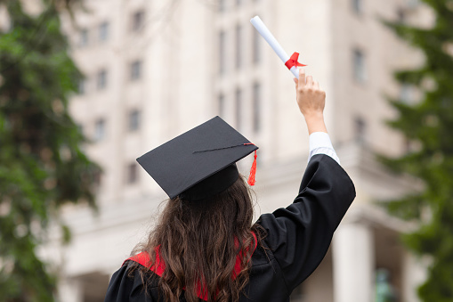 Back view of brunette lady in graduation costume raising her diploma up, unrecognizable female student posing at university campus, celebrating graduation, showing her success, copy space