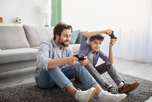 Videogames concept. Joyful father and son competing in online games, holding joysticks while sitting on carpet in living room and spending time together on weekend, copy space