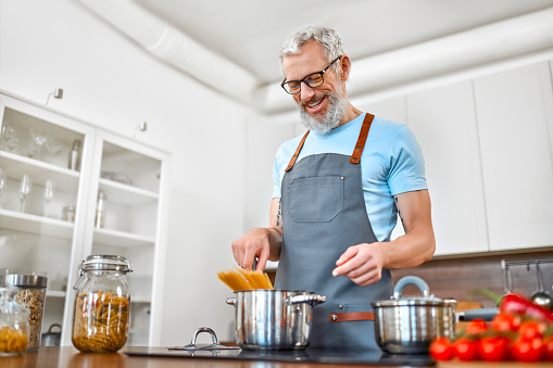 A mature elderly man in an apron prepares pasta in the kitchen. Vegan, vegetarian, healthy lifestyle.