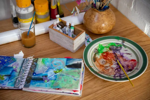 A medium, high angle view of a sketch book and paint on a workbench within a home art studio.