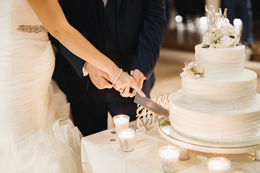 Bride and groom cutting three-tiered cake in white frosting together at a wedding party, close-up . High quality photo