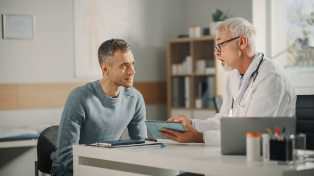 Experienced Middle Aged Family Doctor Showing Analysis Results on Tablet Computer to Male Patient During Consultation in a Health Clinic. Physician Sitting Behind a Desk in Hospital Office. Experienced Middle Aged Family Doctor Showing Analysis Results on Tablet Computer to Male Patient During Consultation in a Health Clinic. Physician Sitting Behind a Desk in Hospital Office. doctor patient stock pictures, royalty-free photos & images