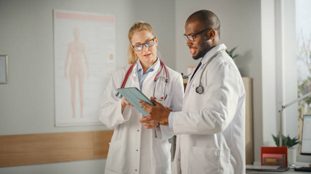 istock Doctor's Office: Black Physician Talks With Professional Head Nurse, Using Tablet Computer. Medical Health Care Specialists Discussing Test Results, Prescription Medicine, Patient Treatment. 1315185781
