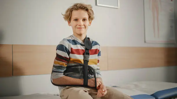 Portrait of a Healthy Young Handsome Teenage Boy Sitting on a Bench with His Hand in an Arm Brace in a Health Clinic. Child in Colorful Jumper is Happy and Smiling in Hospital Office.
