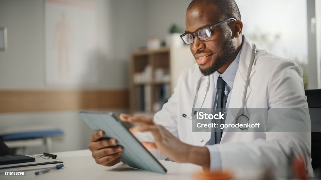 Happy and Smiling African American Male Doctor Wearing White Coat Working on Tablet Computer at His Office. Medical Health Care Professional Working with Test Results, Patient Treatment Planning. Doctor Stock Photo