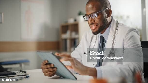 Happy And Smiling African American Male Doctor Wearing White Coat Working On Tablet Computer At His Office Medical Health Care Professional Working With Test Results Patient Treatment Planning Stock Photo - Download Image Now