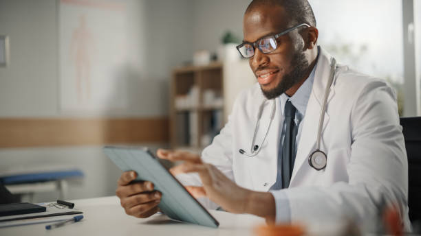 快樂和微笑的非洲裔美國男醫生穿著白色外套在他的辦公室平板電腦上工作。醫療保健專業人員負責檢測結果、患者治療規劃。 - 醫生 圖片 個照片及圖片檔
