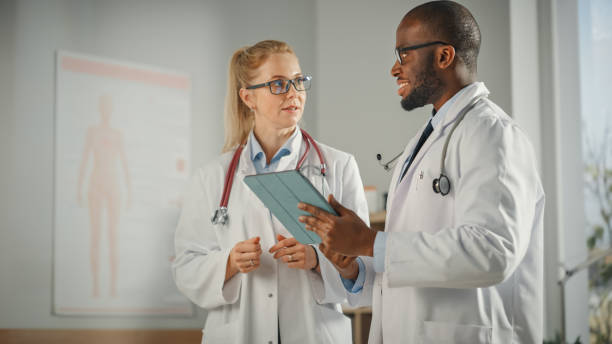 istock Doctor's Office: Black Physician Talks With Professional Head Nurse, Using Tablet Computer. Medical Health Care Specialists Discussing Test Results, Prescription Medicine, Patient Treatment. 1315185818