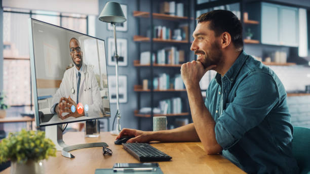 el apuesto hombre caucásico está haciendo una videollamada a su consultor médico en la computadora de escritorio en la sala de estar del hogar mientras está sentado en la mesa. hombre trabajando desde casa y hablando con un médico a través de interne - patient room fotografías e imágenes de stock