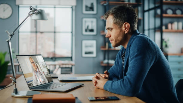 istock Handsome Caucasian Man Having a Video Call with Female Doctor on Laptop Computer while Sitting Behind Desk in Living Room. Freelancer Working From Home and Talking a Medic Over the Internet. 1315185893