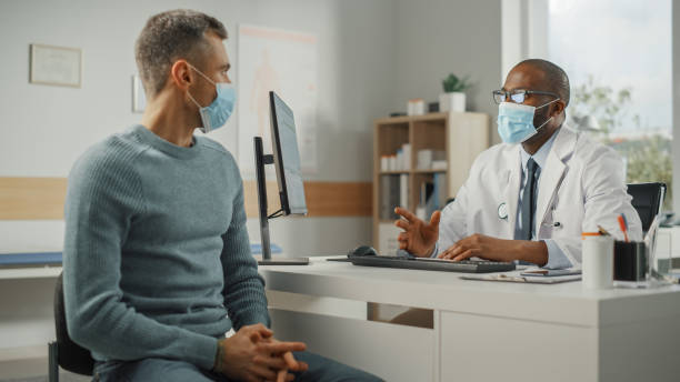 Middle Aged Family Doctor is Talking with Young Male Patient During Consultation in a Health Clinic. Both Wear Face Masks. Physician in Lab Coat Sitting Behind a Computer Desk in Hospital Office. el médico de familia de mediana edad está hablando con un paciente masculino joven durante la consulta en una clínica de salud. ambos usan máscaras faciales. médico con bata de laboratorio sentado detrás de un escritorio de computadora en la oficina