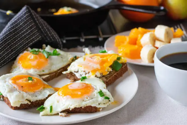 Delicious and savory breakfast with open faced fried egg sandwich served with a plate of fresh fruits and a cup of coffee on a kitchen table at home with blurred background.