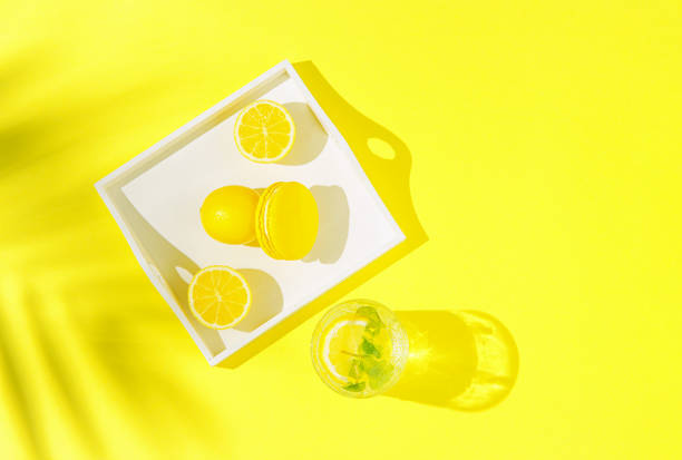 Yellow macaroon with lemons on white tray and glass of lemonade on background with palm tree shadow. Summer refreshment concept. Top view.