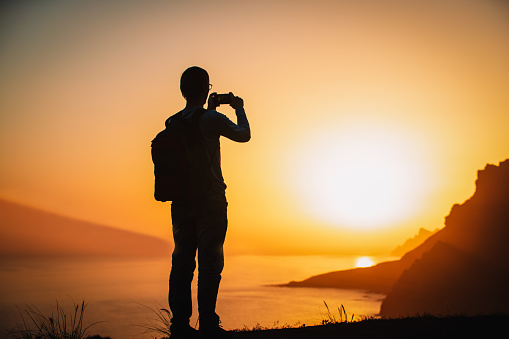 silhouette of a man holding a cellphone taking pictures outside during sunset in mountains. travel concept