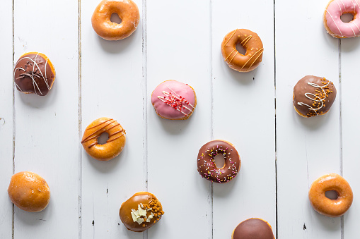 High angle overhead view of a selection of fresh donuts with different colored toppings and glazes. The donuts are arranged on a white wooden surface with room for copy space.