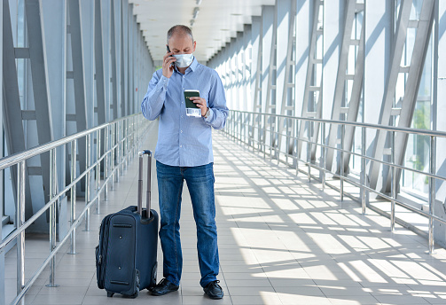 A businessman in a medical mask, with a passport, ticket, suitcase, calls while at the airport or shopping center during the outbreak of coronavirus. Lokdanu, quarantine, flight cancellation