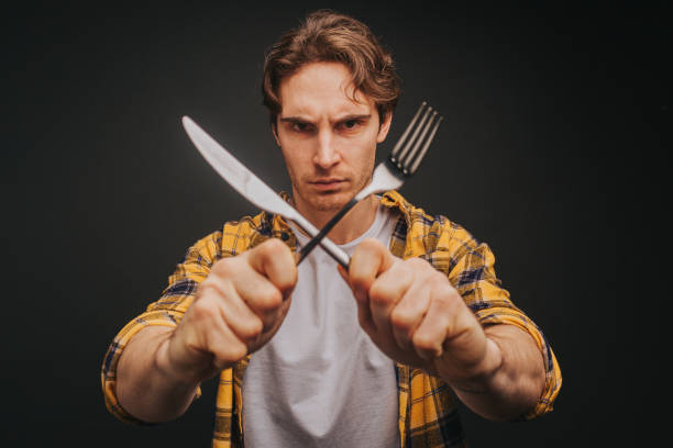 istock Young blond man in yellow shirt is smiling while holding fork and knife making stop, isolated over black background 1315208945