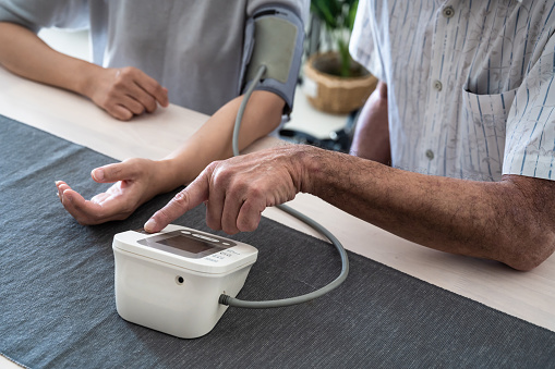 Close-up of an elderly Asian man helping his wife to check her blood pressure and heart rate with digital pressure gauge on desk. Home Medical & Healthcare, Active & Healthy Lifestyle, Medicare & Medical Insurance, Stay at Home Concepts.