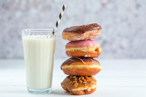 Close up color image of a selection of fresh donuts with different colored toppings and glazes. The donuts are stacked on top of each other on a white wooden surface with room for copy space. There is a glass of milk with the donuts with a drinking straw.