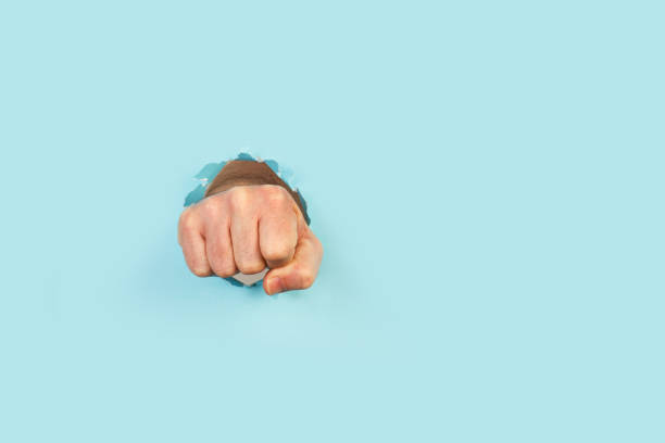 istock Man hand with clenched fist through a hole in a light blue background 1315221870