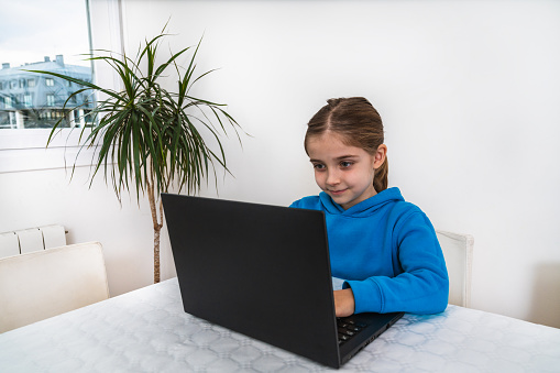 Little blonde girl at home dressed in school uniform with blue sweatshirt studying home school homework with laptop