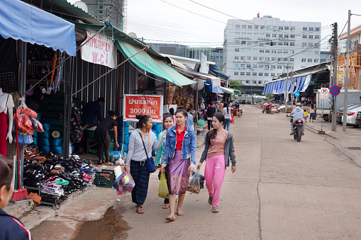 Young laotian women with shopping bags are coming from Khin Din Market in Vientiane. Along street are first market stalls. Main market is in far background area.