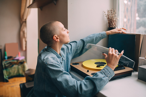 Waist up portrait view of the stylish woman with short hair wearing jeans shirt sitting near her vinyl music player and preparing to listening music. Stock photo