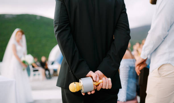 The host at a wedding party stands and holds a microphone behind his back, close-up
