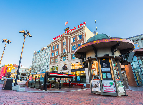 Cambridge, Massachusetts, USA - April 27, 2021: View of Harvard Square as sunet approaches. Visible are the MBTA Harvard Bus and Train station entrance, Cambridge Visitor's Information Center kiosk, and businesses along Massachusetts Avenue. Low, wide-angle view with converging perspective.