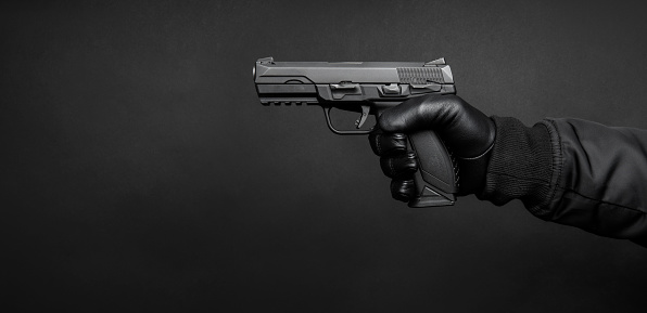 A man's hand in a black leather glove holds a pistol. Weapon in the hand of a man on a dark background. Weapon threat.