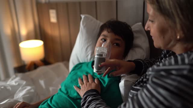 Mother helping son using nebulizer during inhalation therapy in the bed at home