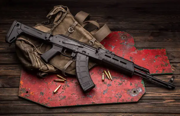 Photo of A classic Soviet military rifle AK in a modern body kit. The carbine and cartridges to it are on near the military backpack and the target for shooting. Weapons on a wooden table.