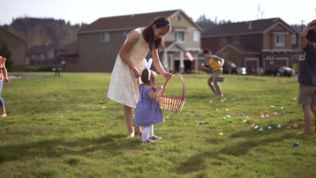 A mixed race pre-adolescent girl affectionately holds her one year old sister's hand and helps her pick up eggs during an Easter egg hunt in their suburban neighborhood park. The sisters are having fun with their friends who are searching for eggs in the background. It's a sunny, warm day and the kids are barefoot.