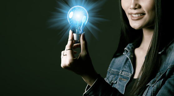 Woman has long straight black hair hold Light Bulb as problem solve and thinking Idea. Portrait of Asian female wear many rings over dark green tone background, copy space