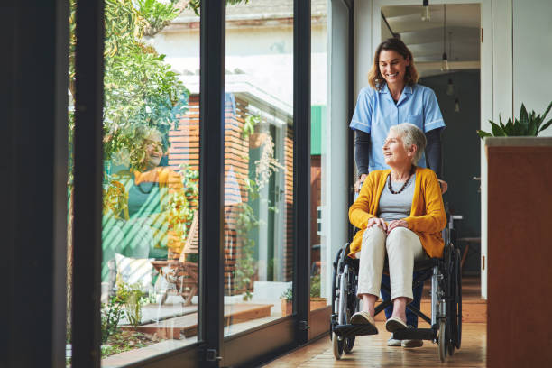 foto de una joven enfermera empujando a una mujer mayor en silla de ruedas en una residencia de ancianos - cuidado fotografías e imágenes de stock