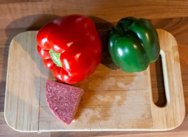 Fresh washed red and green sweet peppers with wedge of spicy salami on a wooden cutting board ready for Italian or Mediterranean cooking in a high angle view