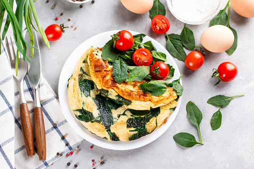 Delicious omelet with spinach, cheese and tomatoes on a white plate on a light slate, stone or concrete background. Top view. Copy space.