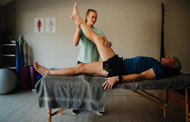 female physiotherapist stretching hamstring of elderly man lying on massage bed in exercise room - bending knees imagens e fotografias de stock