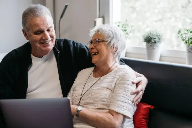 Elderly couple sitting on a sofa, laughing and communicating via laptop